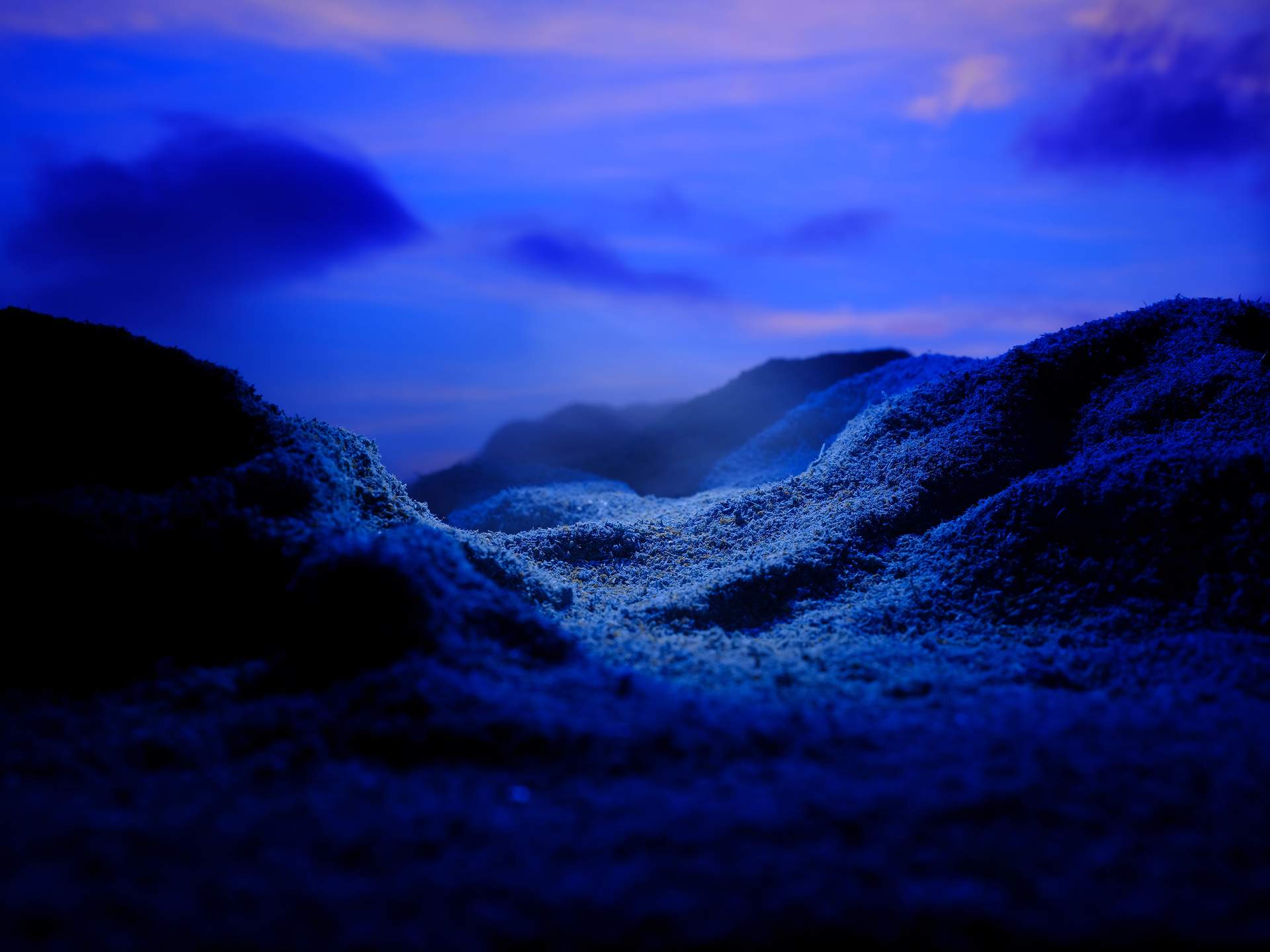 An image showing mountains in a valley, set against a rich blue sky with soft clouds. The centre of this landscape is highlighted in light-blue light, while the rest of the landscape is shrouded in shadow.