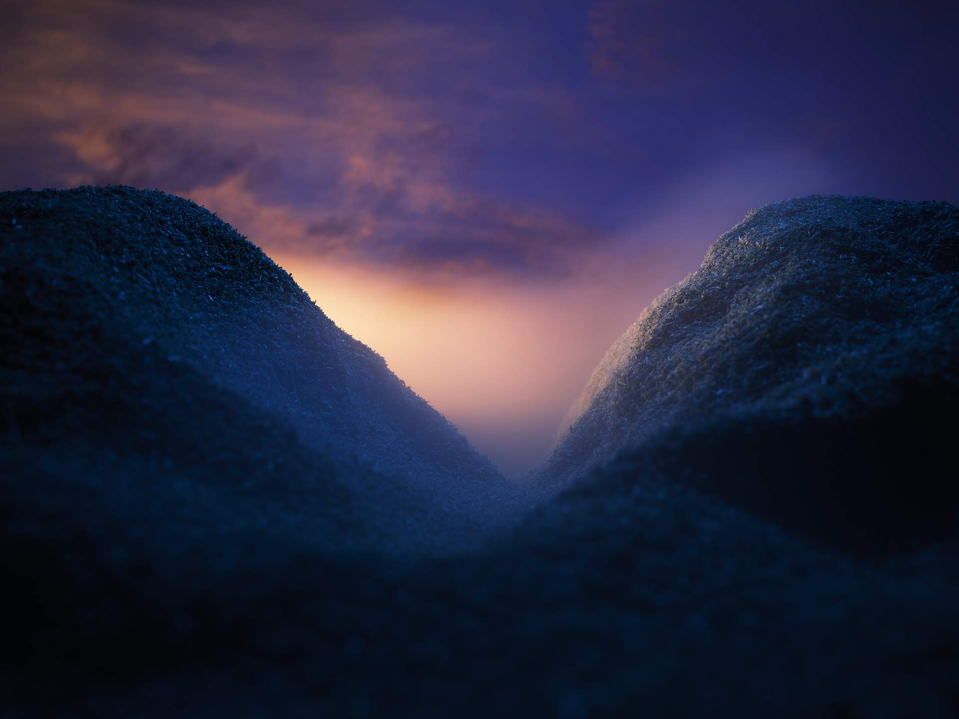 An image of a valley with white-yellow mist emerging from behind a mountain on the left. The mountains in the foreground are shrouded in dark blue shadow, while the sky in the background has clouds tinged with the light of a fading sunset.