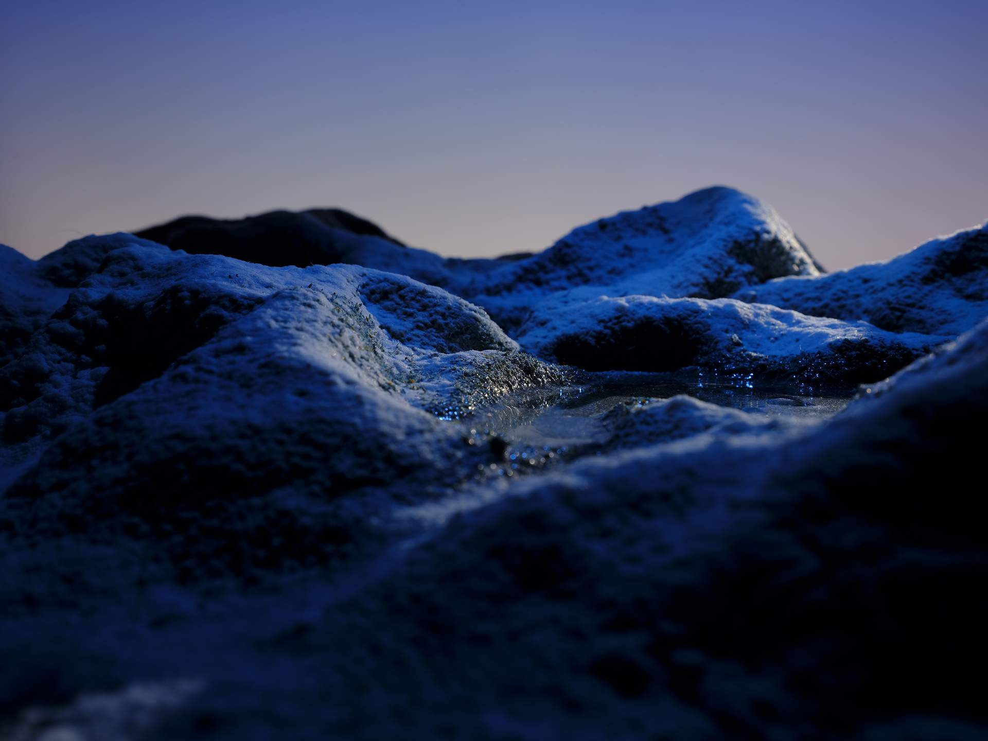 A wintry landscape, covered in snow, can be seen. In the foreground is a small lake, and beyond this are mountains. The gradient of the sky suggests that it is dawn or dusk.